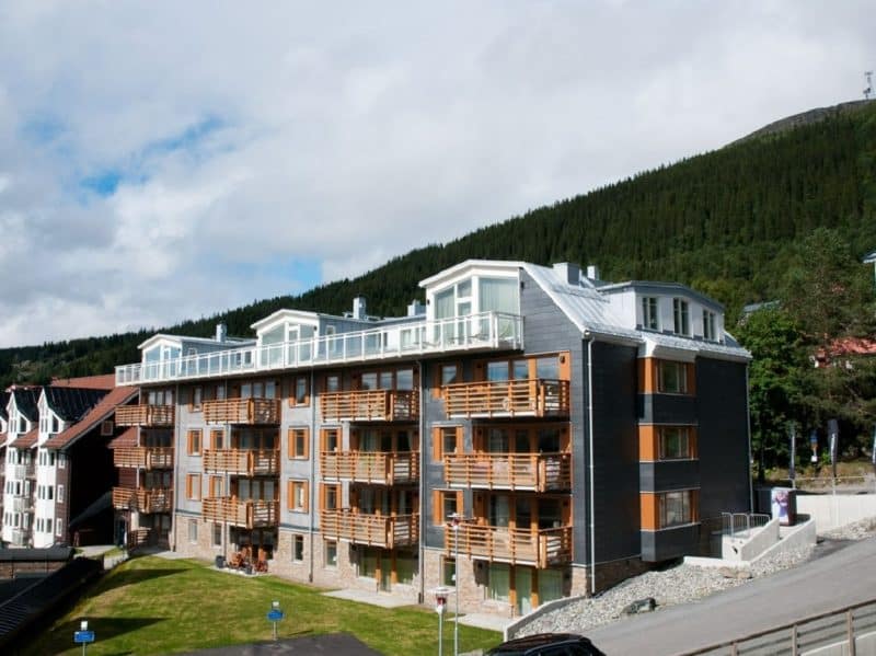 Exterior image of Alpinhuset in Åre. Dark gray property with orange-brown balcony railings and light gray tin roof. In the foreground a green lawn. in the background dark green forest on Åreskutan.