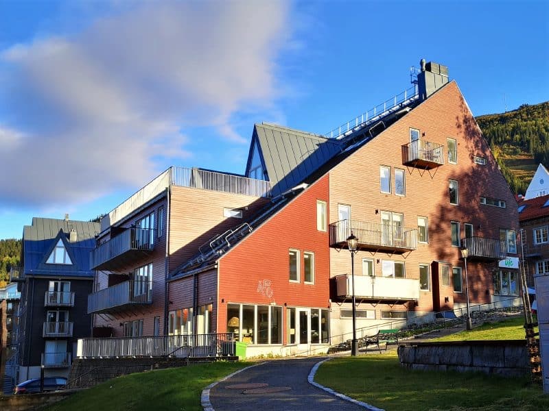 Red-brown modern building with asymmetrical, steeply pitched roof, balconies with different types of railings and restaurant APO on ground level. In front of the property is a gravel path through a park with a green lawn and a stone wall. in the background more properties can be seen as well as Åreskutan and a blue sky with a few clouds.