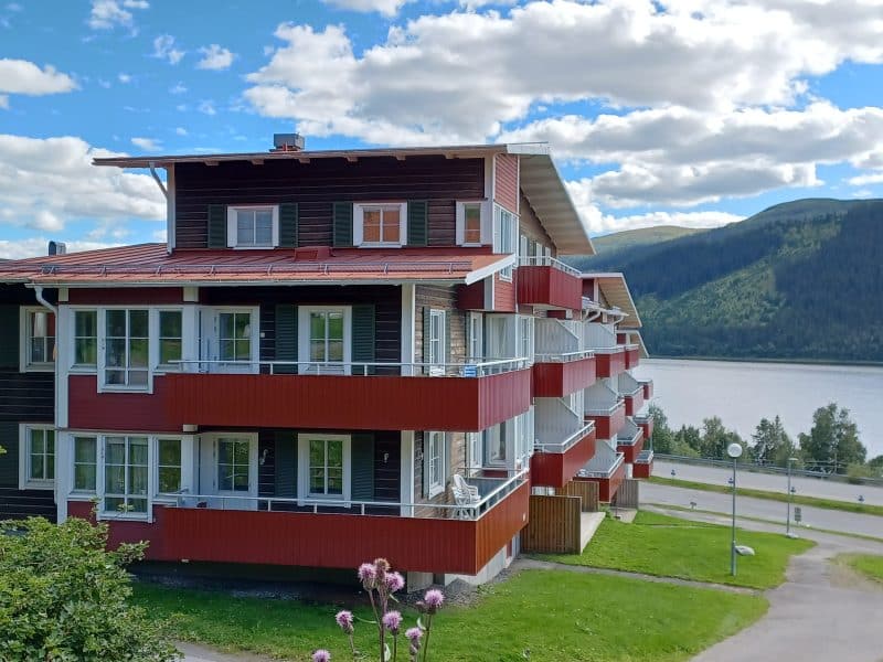 Brown house on three levels with white details and red balcony railings. Located on a green grassy slope. below you can see a road as well as Lake Åresjön, Reinfjället and a blue summer sky with white cumulus clouds.
