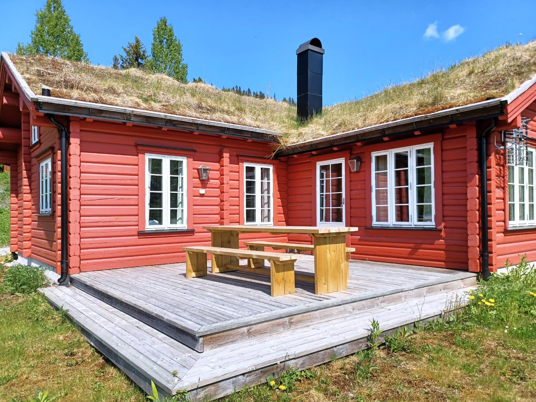 Wooden deck with wooden outdoor furniture at a red log cabin built at an angle. One-story house with sedum roof. A tall black chimney on the roof. White window frames and moldings. Black lanterns as wall lighting. Green lawn and blue summer sky.