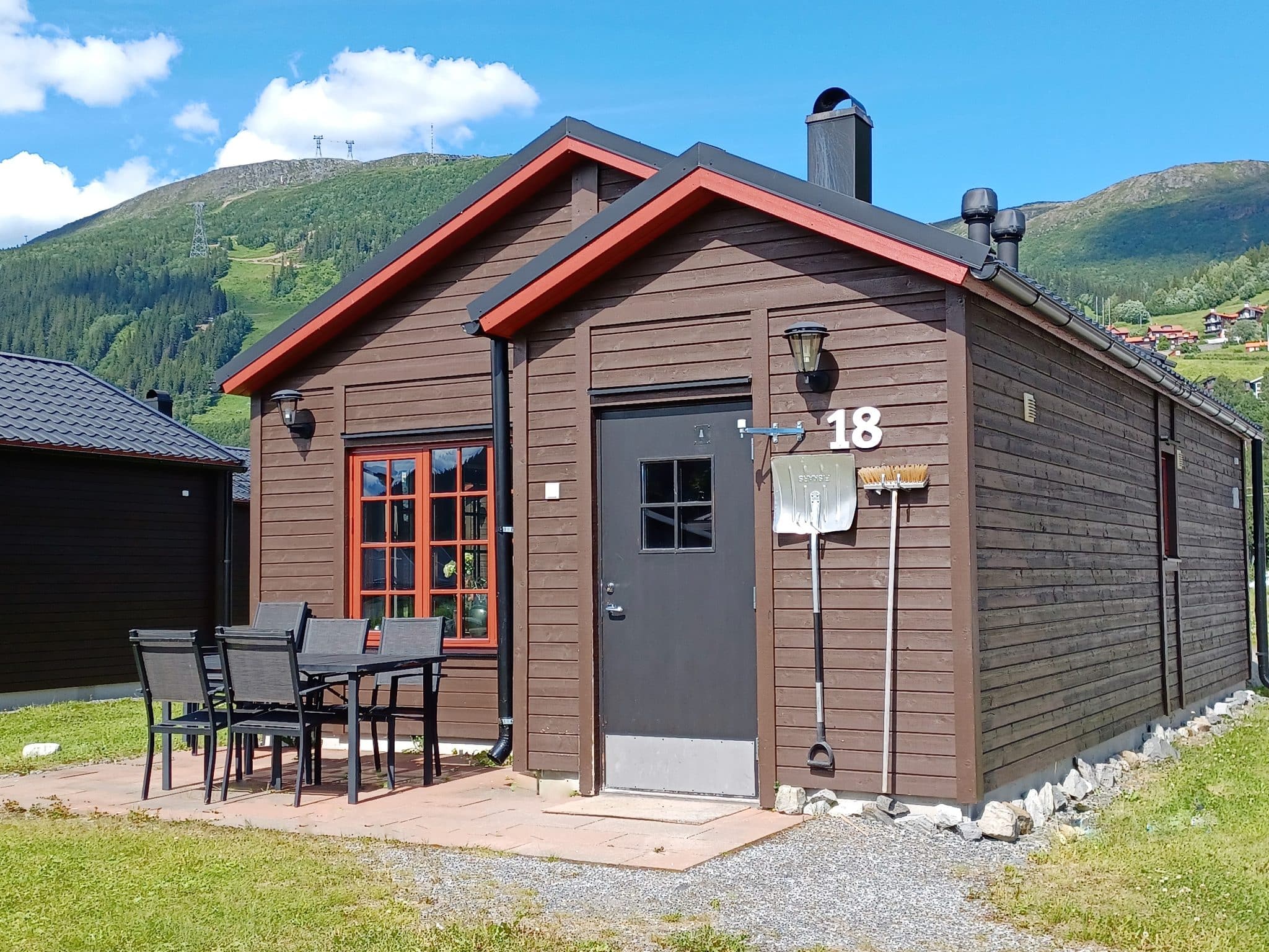 Brunt fristående hus med uteplats med utemöbler. På väggen till höger om den mörkgrå ytterdörren hänger en snöskyffel och en sopkvast. I bakgrunden en grön Åreskuta och en blå sommarhimmel.
