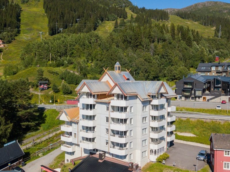 Large white house with 6 floors where all apartments have balconies and a lower floor. In the foreground you can see the roof of the Broken restaurant. Behind you can see the lift up to the VM6, verdant ski slopes and other accommodation.