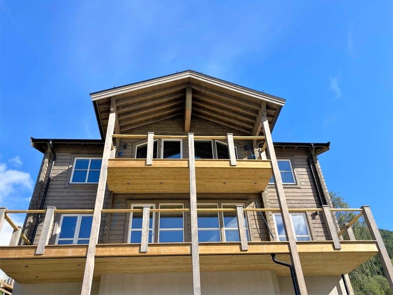 Picture taken in so-called frog perspective of a light gray log house on three levels with a large balcony on the first floor and a smaller balcony on the second floor. in the background you can see green trees and a blue summer sky.