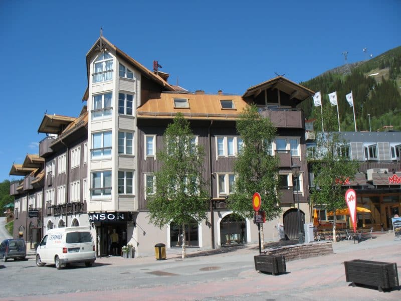 Brown-white property at Åre square in four floors. The Hansson store is located on the ground floor. On the right is the fast food chain Max. In the background you can see a verdant Åreskuta and a blue summer sky.