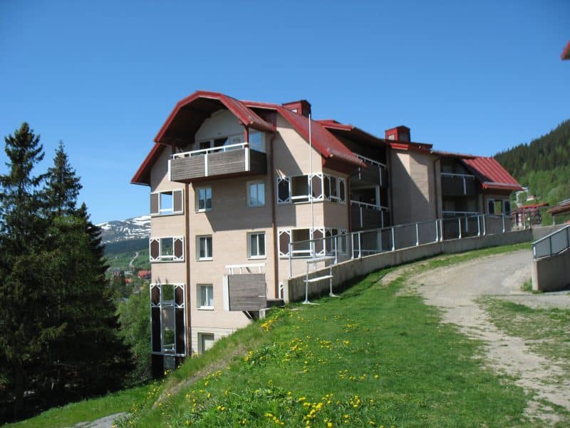 Summer exterior with green grass and blue summer sky of Mitt i Åre 3, a beige-colored apartment building with a red tin roof, five floors in suterra, right next to the ski bridge from the slopes by the cable car.