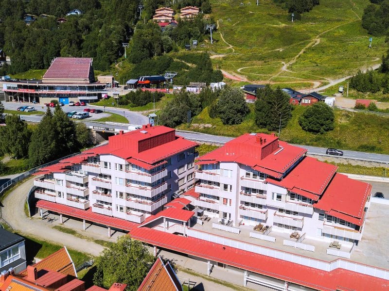 Drone photo of the property in the middle of Åre 1. A light gray apartment building with a red tin roof between the cable car and Åre square. In the background you can see the cable car's valley station, Restaurant Timmerstugan and green slopes with cycle trails.