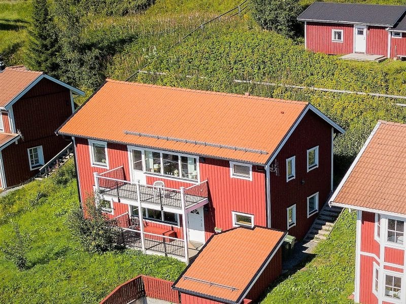Rött tvåplanshus med vita detaljer. Altan och balkongräcke i svart järn och tak med röda tegelpannor. Runt om huset är det gröna gräsmattor och fler röda hus.