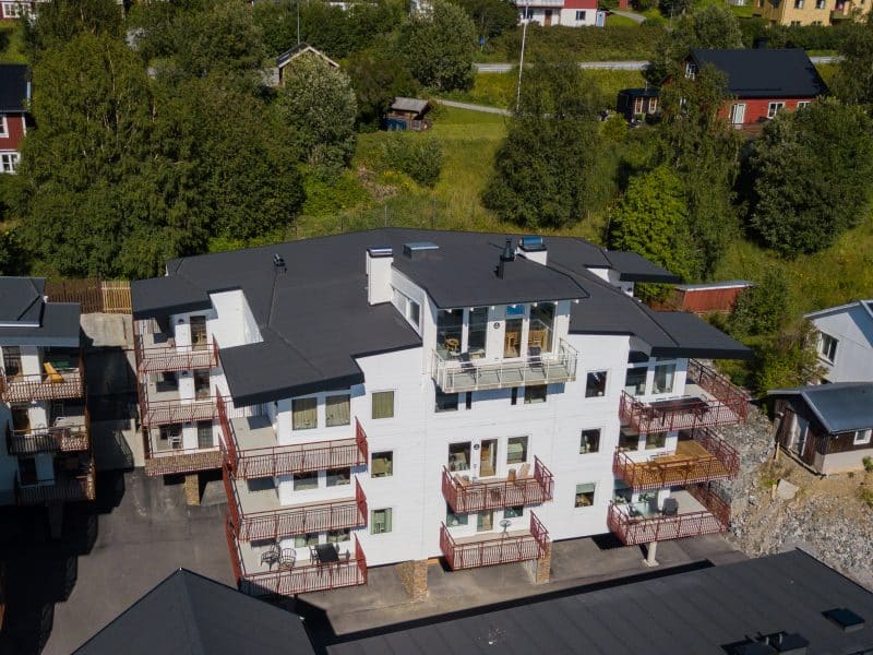 Vitt lägenhetshus med svart plåttak och balkonger. gröna träd och mindre hus i omgivningen runt om.