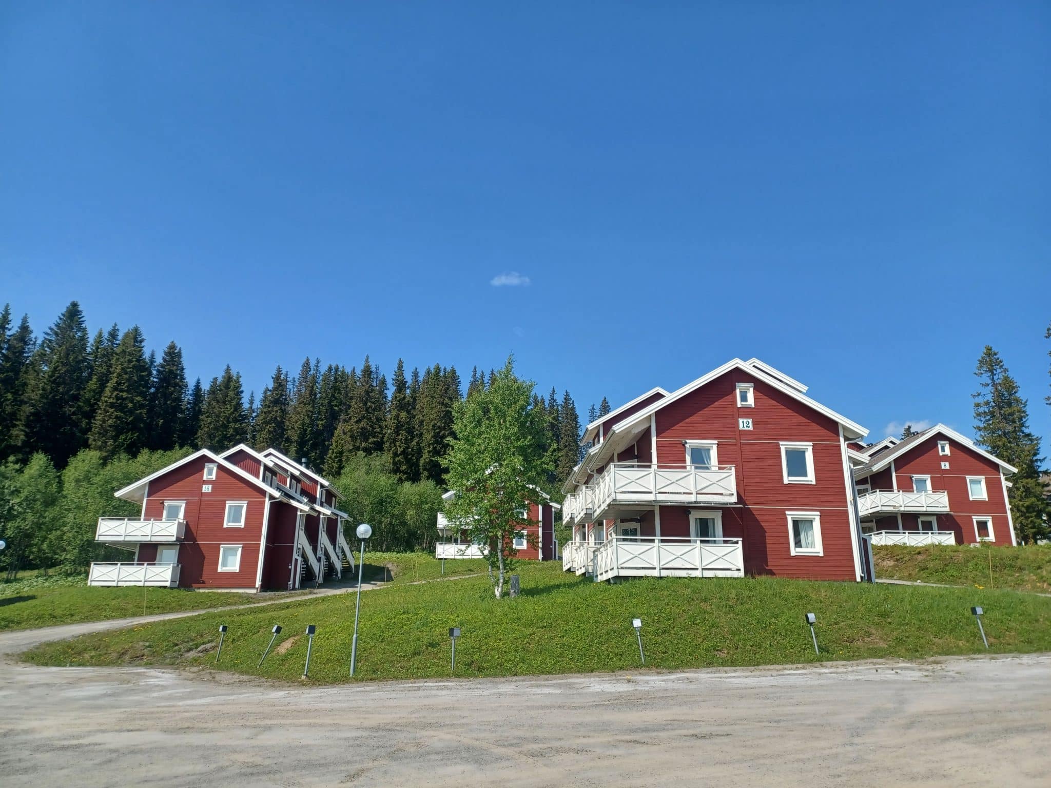 Several red 2 1/2 story houses with white knots, window trim and balcony railings on a large green lawn in front of a green forest and a blue summer sky. in the foreground you can see a gravel field with the property's parking spaces.