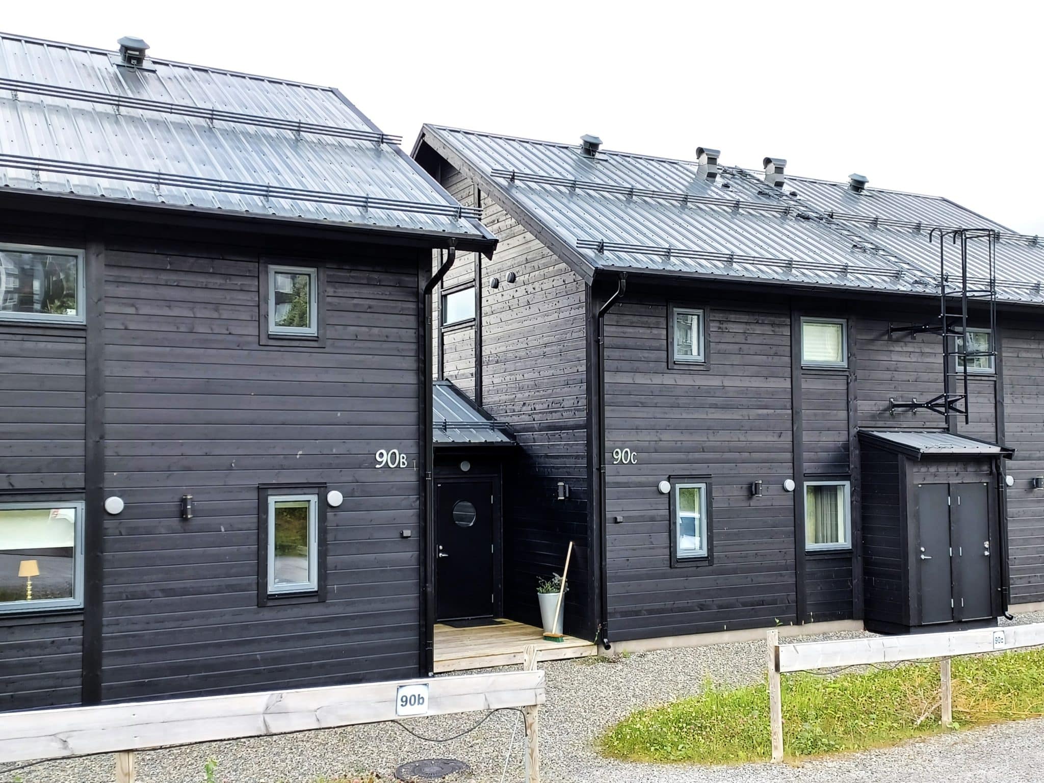 Black semi-detached houses with gray tin roofs on two levels. The houses are tied together by their entrances, which are in a small single-storey extension between each house body. On the facade of the house there is a ski storage room and a fire escape. The ground in front of the house is shingle and there is a small lawn and a low wooden fence separating the property's parking spaces.
