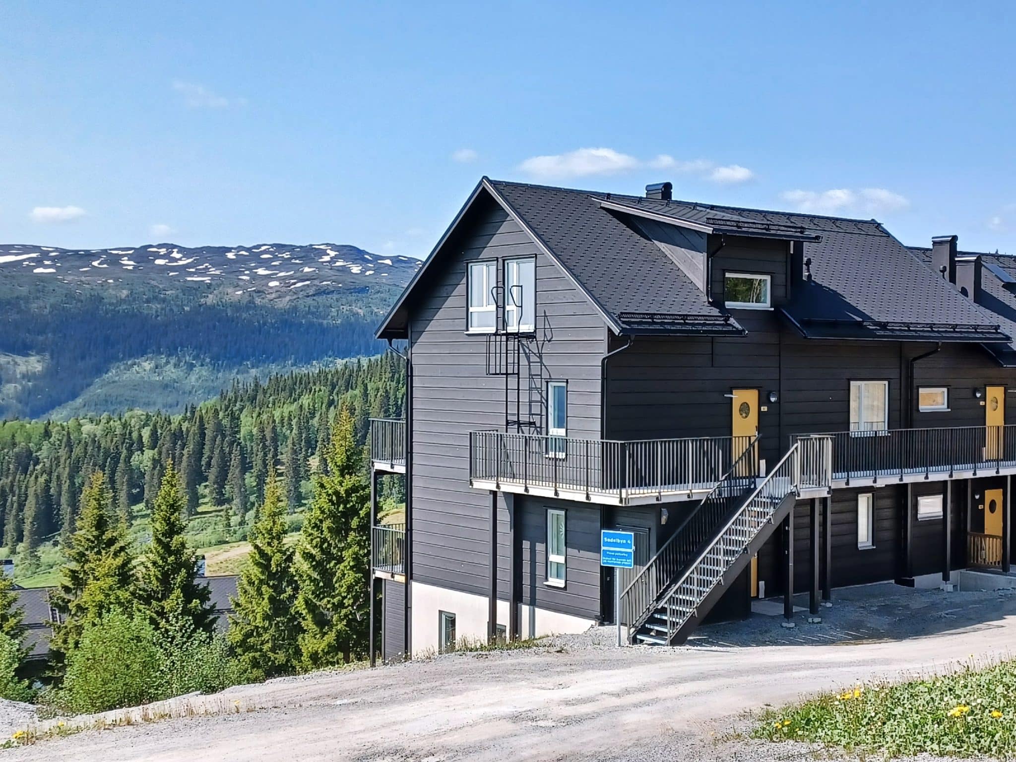 Brown semi-detached property with yellow doors, dormer with windows, external staircase and balcony along the entire facade. In the background a fantastic view of a mountain landscape with patches of snow on top. spruce forest and a blue summer sky.