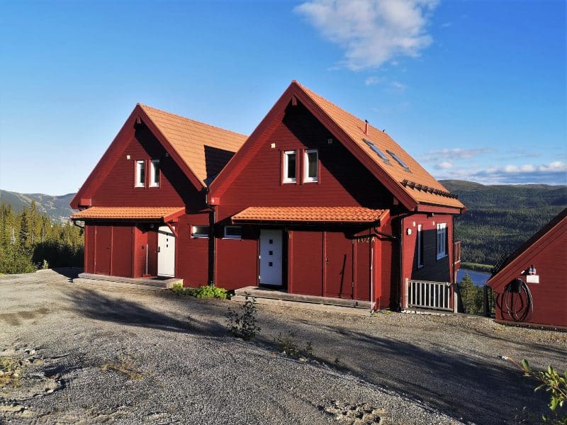 Ett rött parhus i 1 1/2 plan i suterräng med vita dörrar och rött tegeltak. i bakgrunden syns Renfjället och Åresjön.